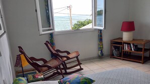 Een slaapkamer met een bed, twee schommelstoelen, een tafeltje met een lamp en uitzicht op zee door een open raam.