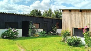 En träbyggnad med en svart dörr och fönster, omgiven av en välskött trädgård med olika växter och blommor.