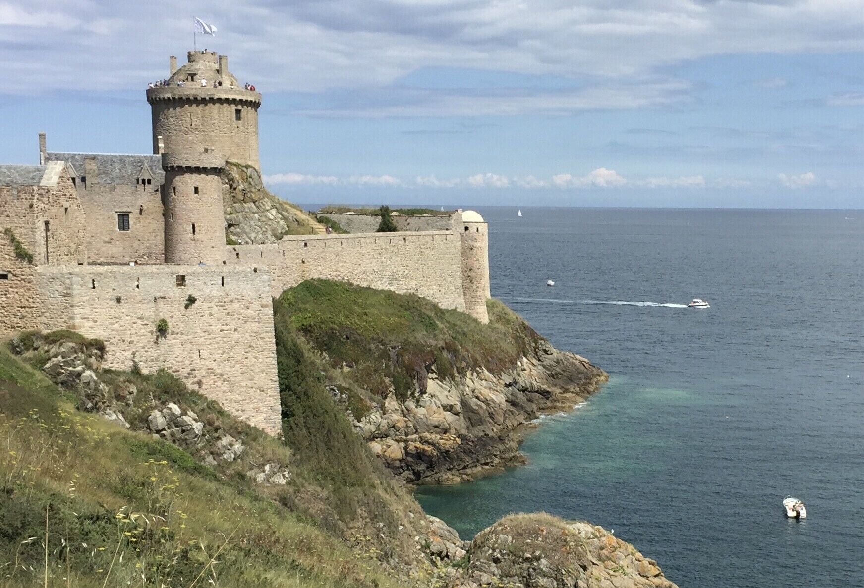 A historic stone castle with a round tower, situated on a rocky coastline with clear blue waters.