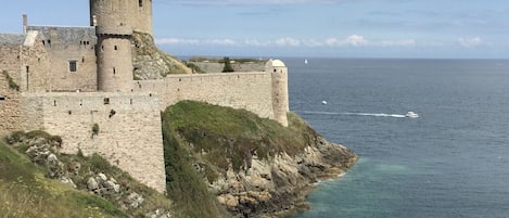 A historic stone castle with a round tower, situated on a rocky coastline with clear blue waters.