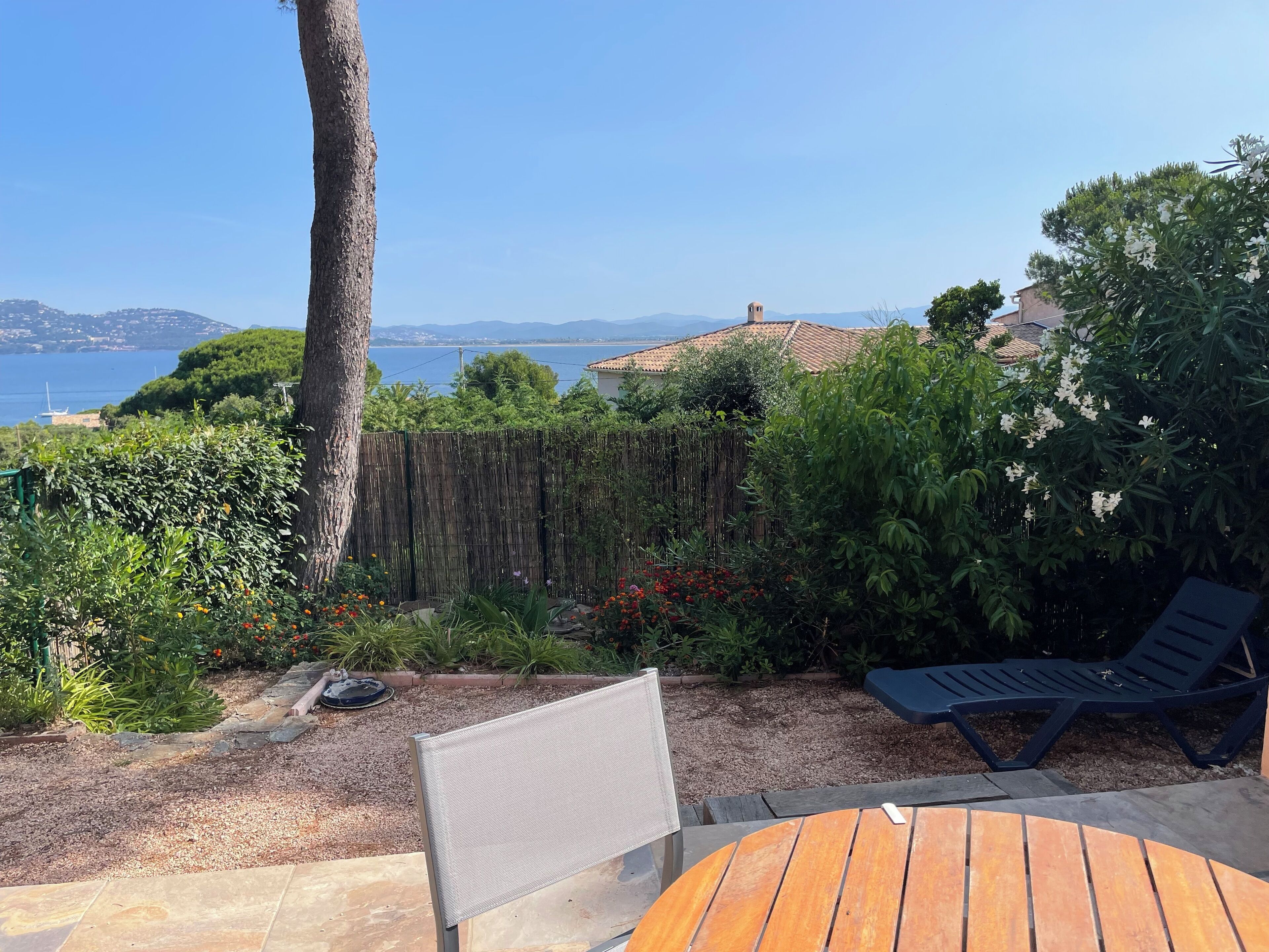 A patio with a wooden table and two lounge chairs, a wooden fence, and a view of the sea and distant hills.