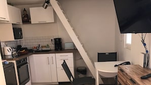 Et kompakt køkken med hvide skabe, en mikroovn, en brødrister og en kaffemaskine. Der er et træspisebord med en stol, og et fjernsyn monteret på væggen.