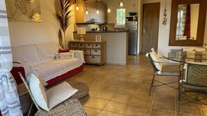 A cozy living room with a red sofa, wicker poufs, and a glass dining table with chairs. A kitchen with wooden cabinets and a refrigerator is visible in the background. - San Cipriano (St Cyprien) 6 pers conditioned villa, in residence (Lecci)