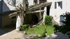 A white building with a tiled roof, a covered entrance, and a small garden area with a tree and flowers.