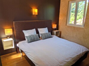 A bedroom with a large bed, two bedside lamps, and a window with a view of greenery.