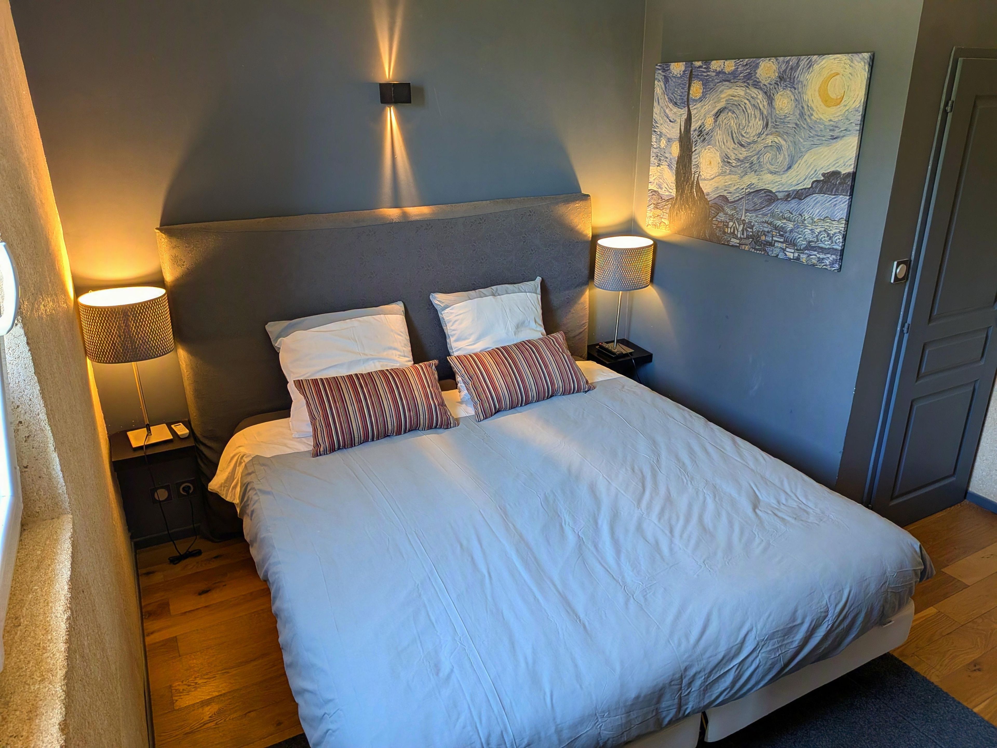 A bedroom with a bed, two bedside lamps, a wall-mounted light, and a painting.