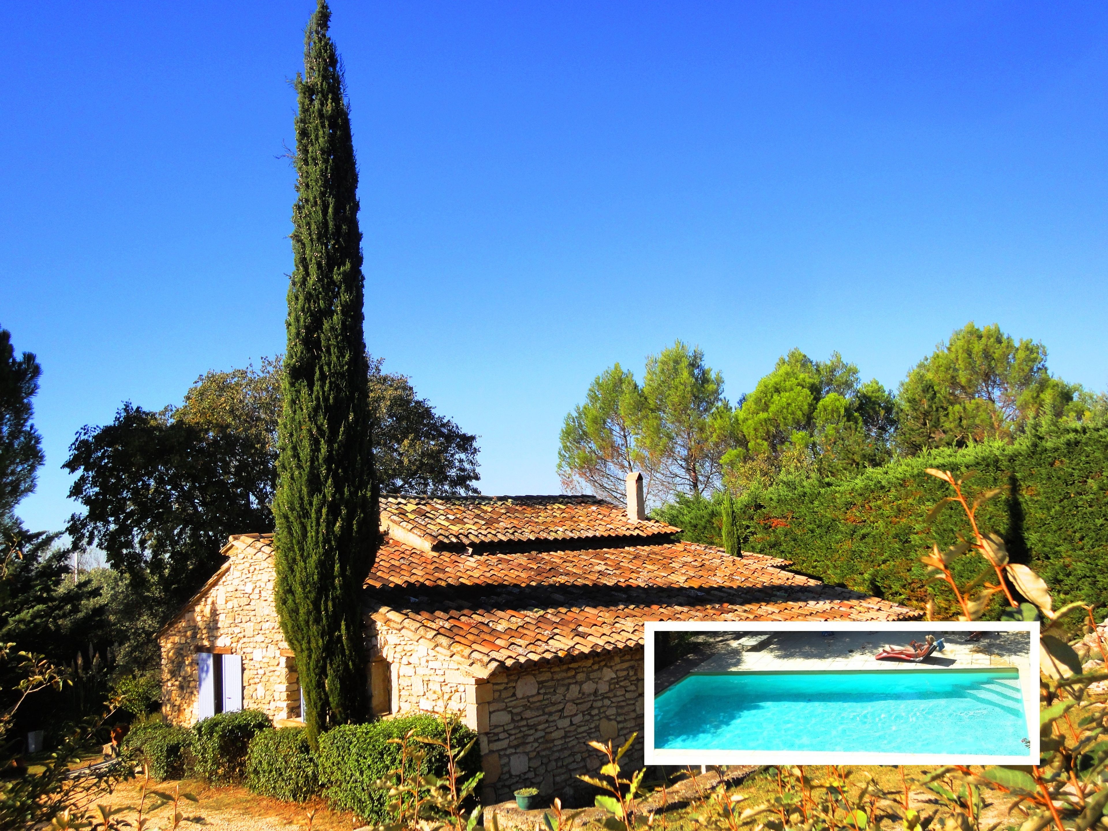 A stone house with a tiled roof, surrounded by greenery and a clear blue sky.