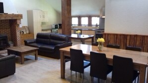 Un salon avec une cheminée, un canapé en cuir noir, une table à manger avec des chaises, et une cuisine en arrière-plan.
