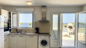 Uma cozinha moderna com máquina de lavar, cafeteira e vista para o mar através de uma grande janela.