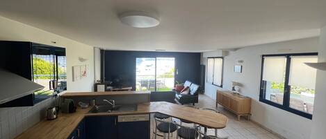 A modern kitchen with a gas stove, a built-in oven, and a dishwasher. There is a dining area with a wooden table and chairs, and a sofa. The kitchen has large windows and a view of the outdoors.
