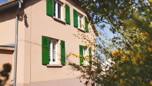 Un bâtiment à la façade beige et aux volets verts, doté de nombreuses fenêtres et d’une cheminée bien visible.