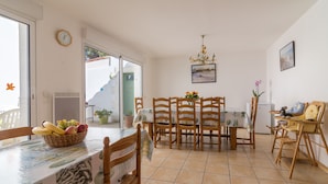 A dining area with a table set for four, surrounded by wooden chairs. There is a basket of fruit on the table, and a small table with a vase of flowers. A clock and a painting are on the walls. - Island of Re. House near sandy beach, harbor and shops (La Flotte)