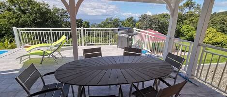 A covered outdoor patio with a round table and chairs, a grill, and a pool in the background.