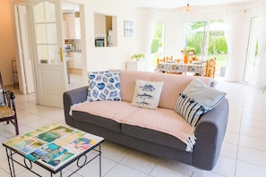 A living room with a grey sofa, patterned cushions, a coffee table with magazines, a kitchen in the background, and a dining area with a table and chairs.