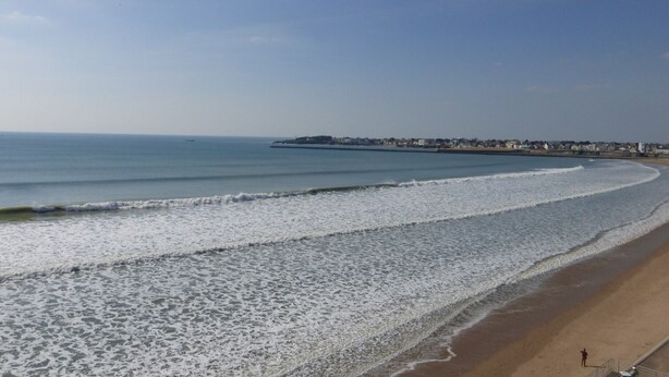 A beach with waves, a pier, and buildings in the distance.
