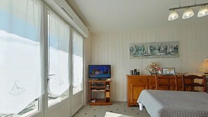 A room with a large window, a television, a wooden cabinet, a bed, and a painting of sailboats.