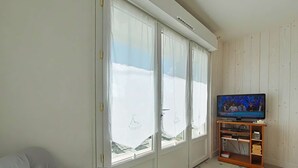 A room with a grey sofa, a television on a wooden stand, and large windows with curtains.