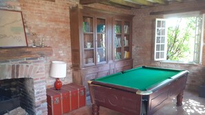 A billiards table in a room with a brick fireplace, wooden cabinets, and a window with a view of greenery. - Gite in Normandy with private and heated swimming pool. (Bacqueville-En-Caux)