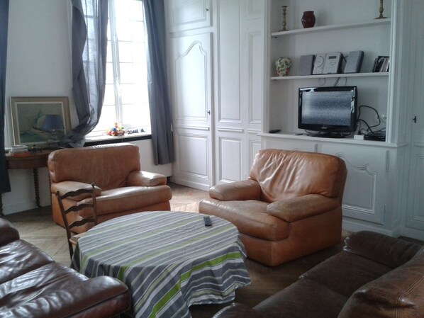 A living room with leather sofas, a striped table, a television on a shelf, and a window with curtains. - Gite in Normandy with private and heated swimming pool. (Bacqueville-En-Caux)