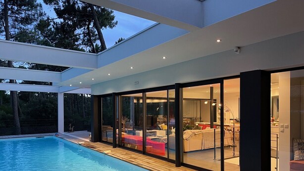 Une maison moderne dotée d’une piscine, de grandes portes vitrées donnant sur un espace de vie et offrant une vue sur l’extérieur.