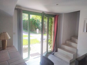 Una habitación con puerta corrediza de vidrio que da a un jardín, una mesita con lámpara y una escalera.