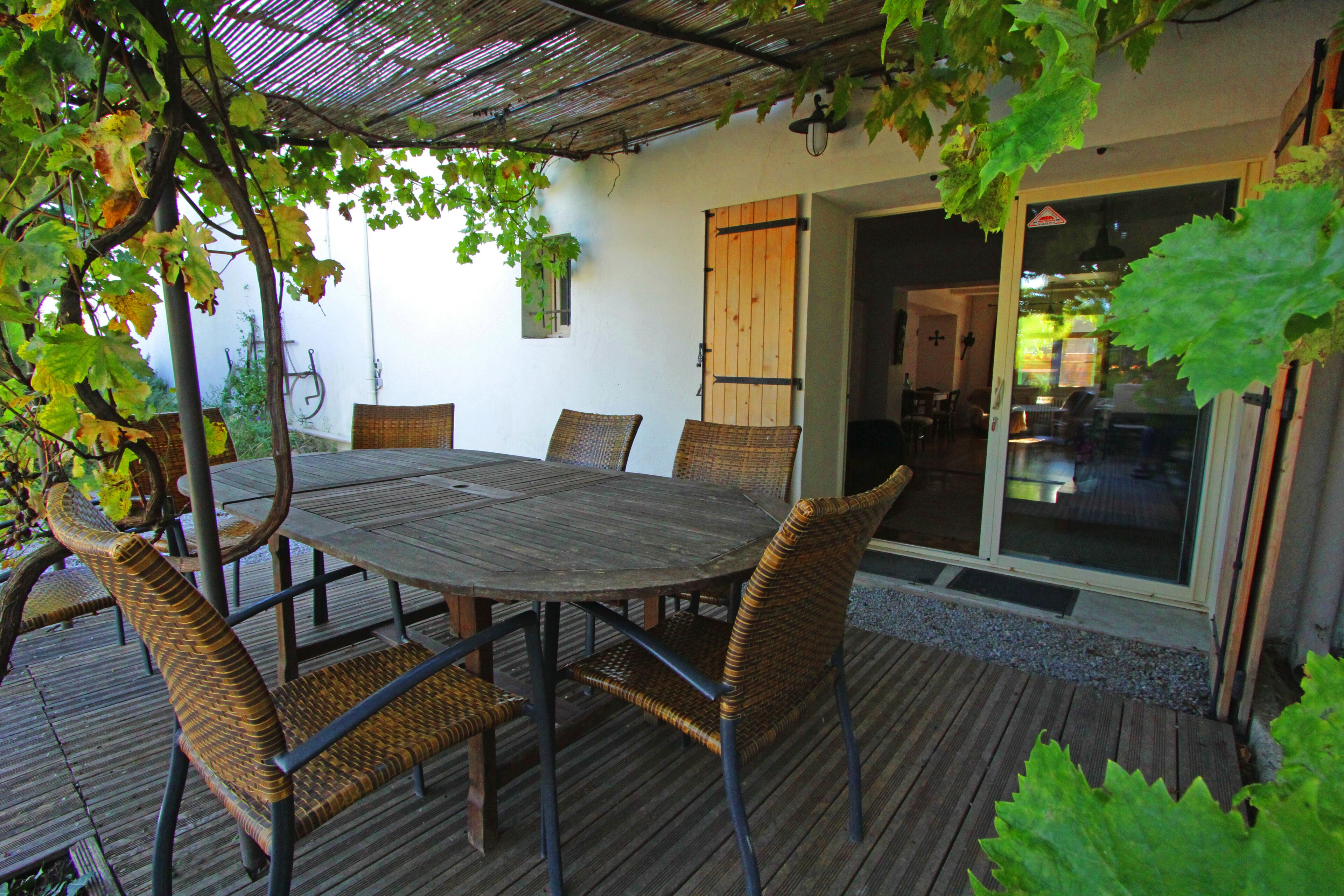 An outdoor patio with a round wooden table and wicker chairs, surrounded by greenery and a pergola.