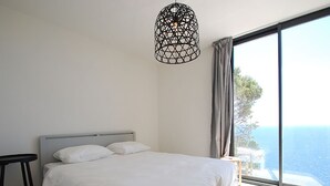 Quarto com cama grande, mesa de cabeceira, luminária pendente e vista para o mar.