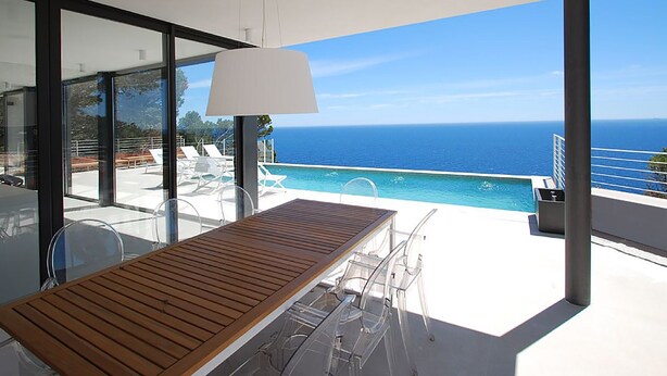 Espaço de refeições moderno ao ar livre, com mesa e cadeiras de madeira, com vista para uma piscina e o mar.