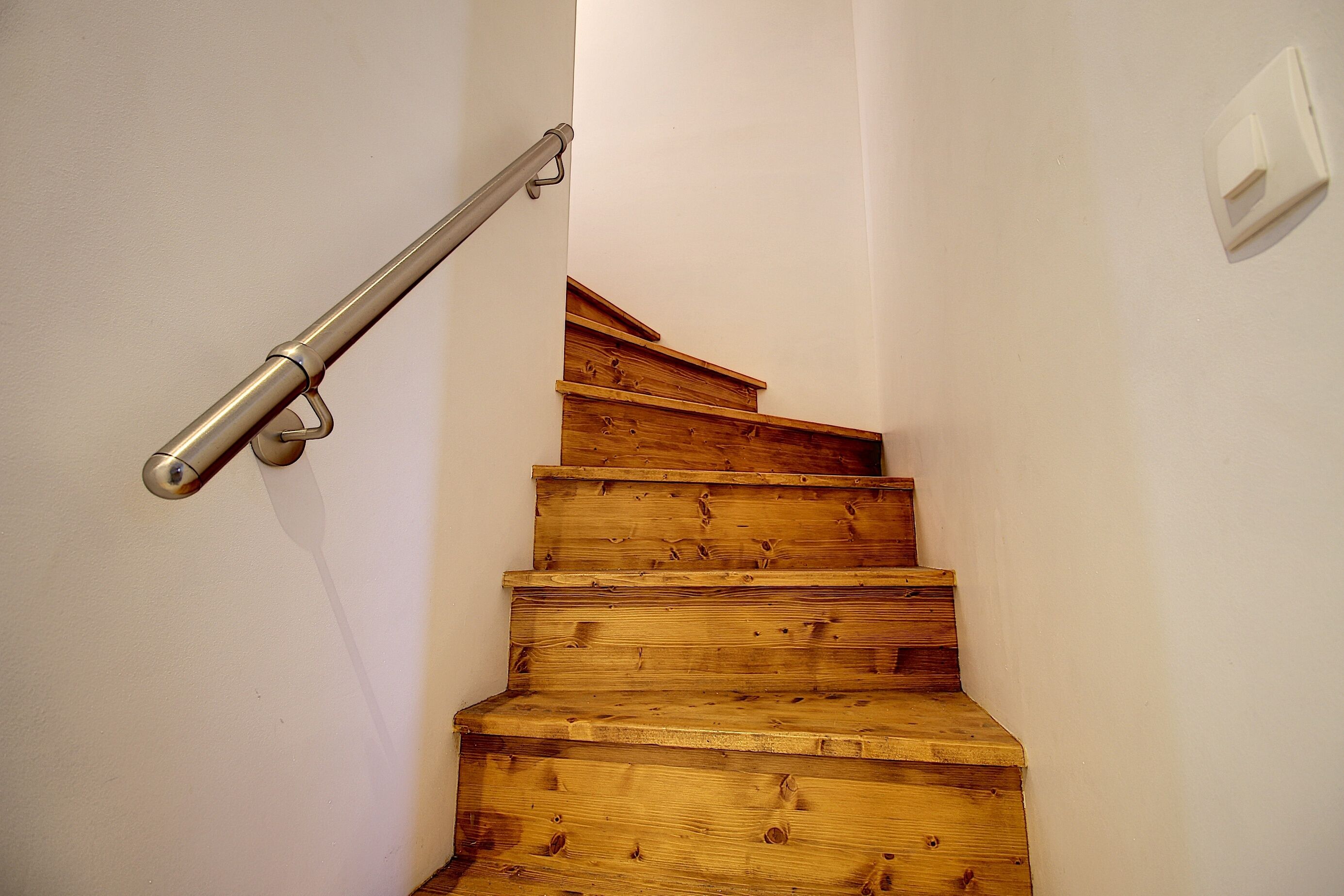 Una scala in legno con corrimano in metallo, pareti bianche e un interruttore a muro.