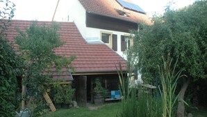 Una casa con tetto spiovente rosso, un camino ben visibile e un giardino con diverse piante e una recinzione in legno.