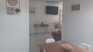 Un coin repas avec une table en bois et des chaises blanches, un téléviseur fixé au mur et une horloge sur le côté gauche.