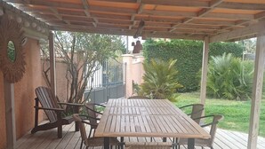 Une terrasse extérieure couverte, avec une table et des chaises en bois, entourée d’une terrasse en bois et offrant une vue sur le jardin.