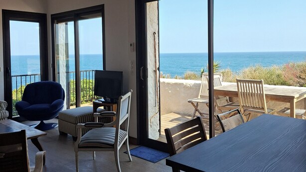 A room with a large sliding glass door leading to a balcony with a view of the sea. Inside, there is a dining table, chairs, a TV, and a blue chair. - Renovated villa with exceptional panoramic view (Banyuls-Sur-Mer)
