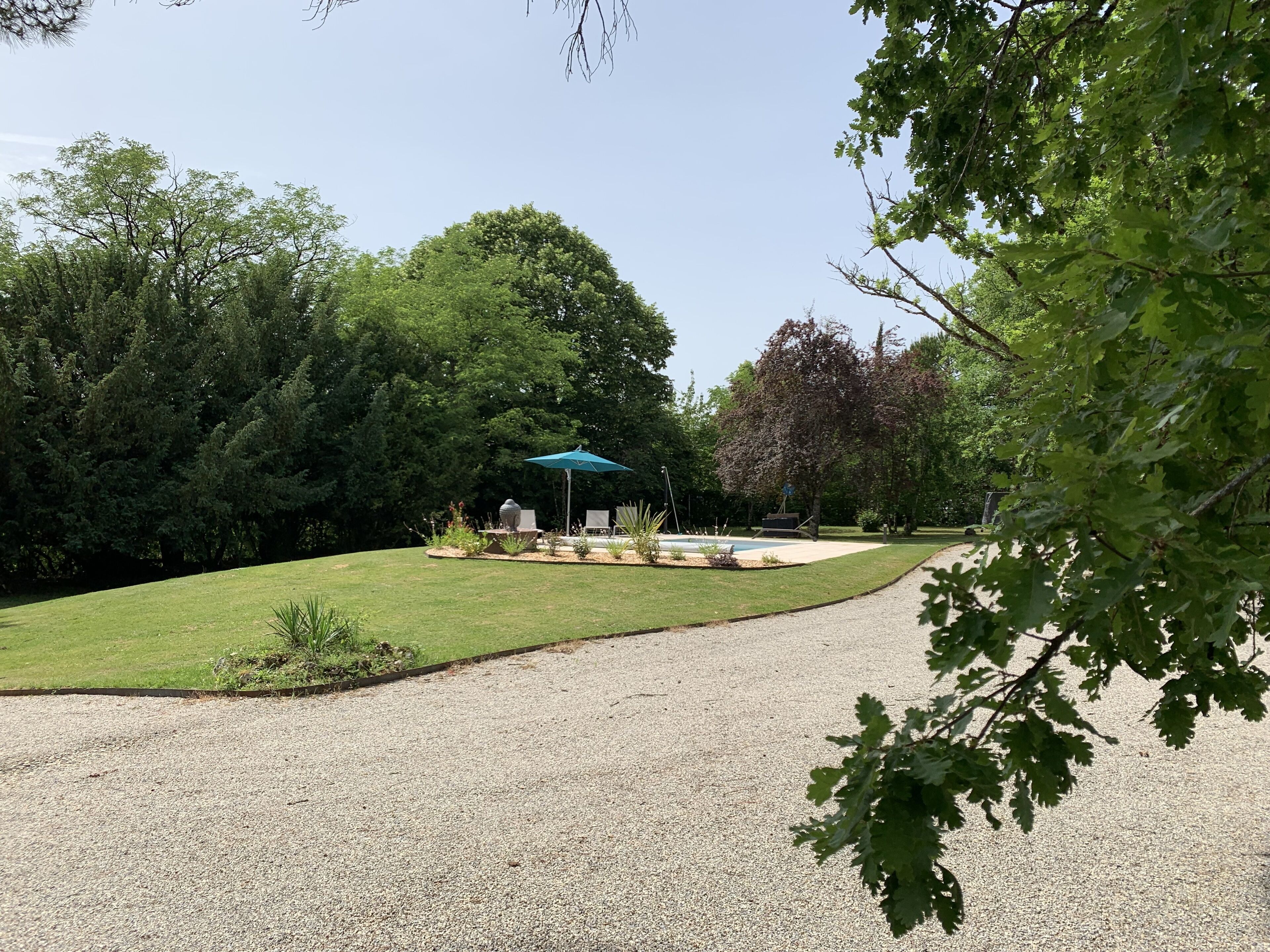 Une allée de gravier mène à un patio agrémenté d’un parasol bleu, le tout entouré d’une végétation luxuriante et d’arbres.