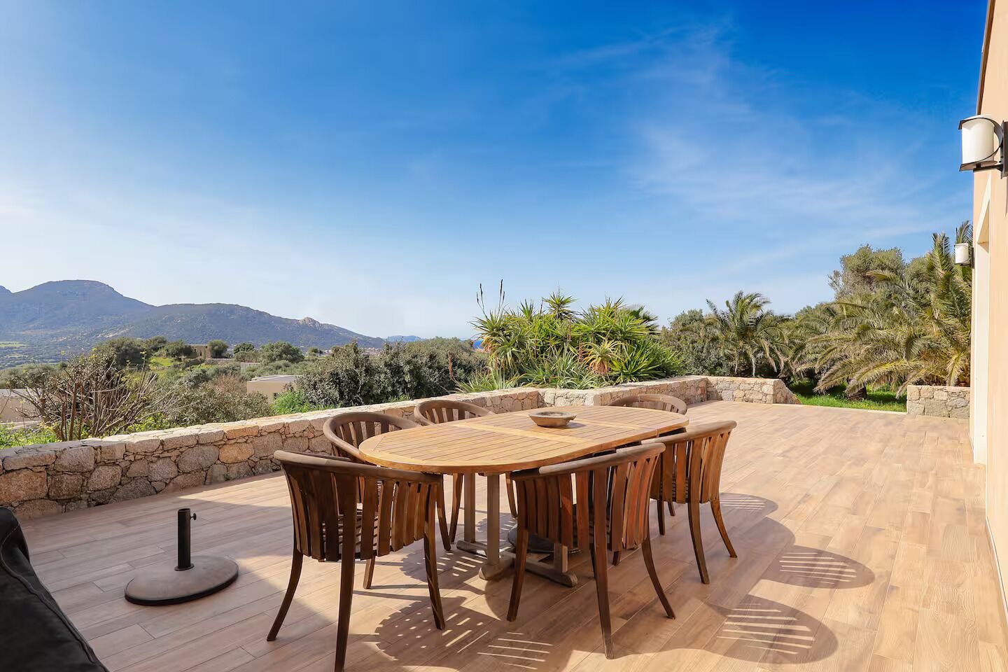 Une table et des chaises en bois sur une terrasse extérieure, adossées à un mur de pierre, avec vue sur un paysage montagneux.