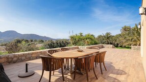 A wooden table and chairs on an outdoor patio with a stone wall, overlooking a mountainous landscape.