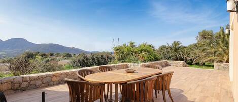 Mesa y sillas de madera en un patio al aire libre con un muro de piedra, con vista a un paisaje montañoso.