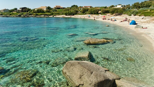 A beach with clear water, rocks, and buildings in the background.