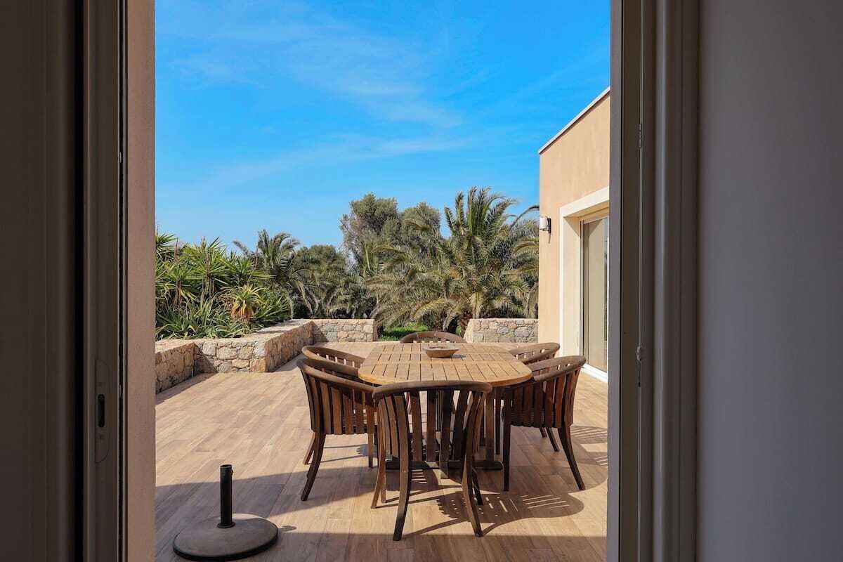 A patio with a wooden table and chairs, surrounded by a stone wall and palm trees.