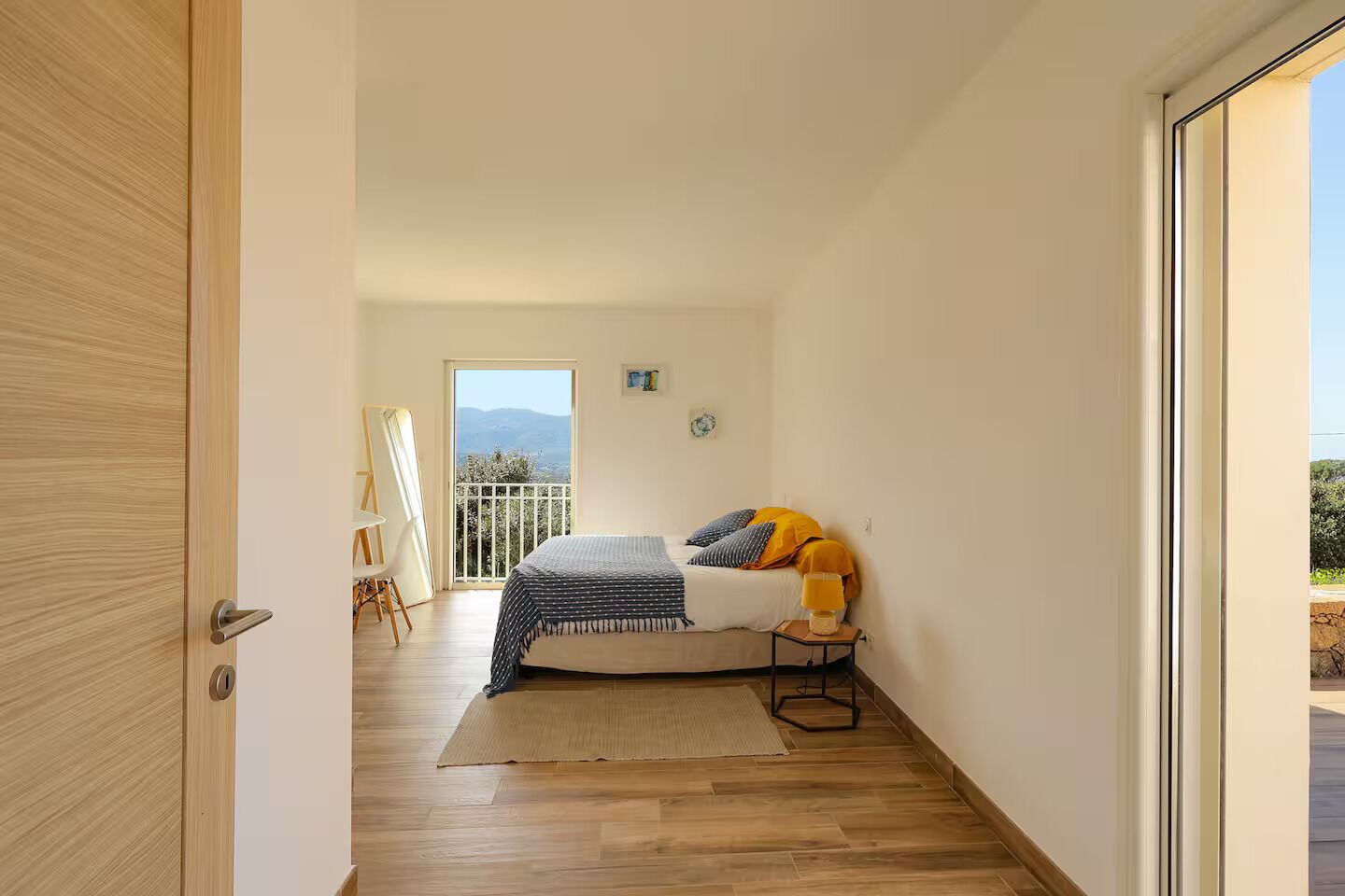 A bedroom with a bed, a small table, a chair, and a balcony with a view.