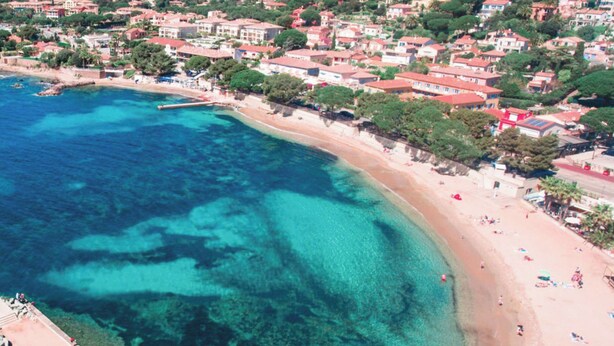 An aerial view of a coastal area with a beach, clear blue waters, and a densely built residential area with red-roofed houses.