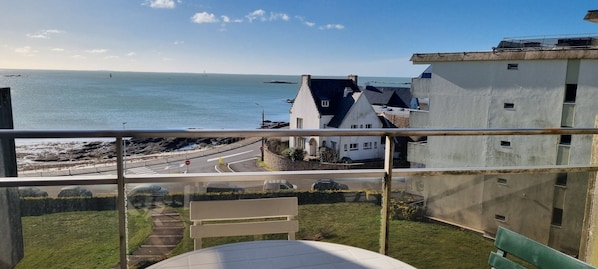 A balcony with a view of the sea, a road with vehicles, and buildings in the background.