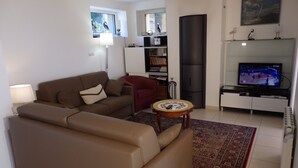 A living room with a brown sofa, a red armchair, a small round table, a bookshelf, a TV, and a large window. - Modern house with garden and beautiful sea view (Pléneuf-Val-André)