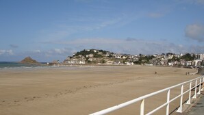 A sandy beach with a white railing, a rocky hill in the background, and a residential area with houses. - Modern house with garden and beautiful sea view (Pléneuf-Val-André)
