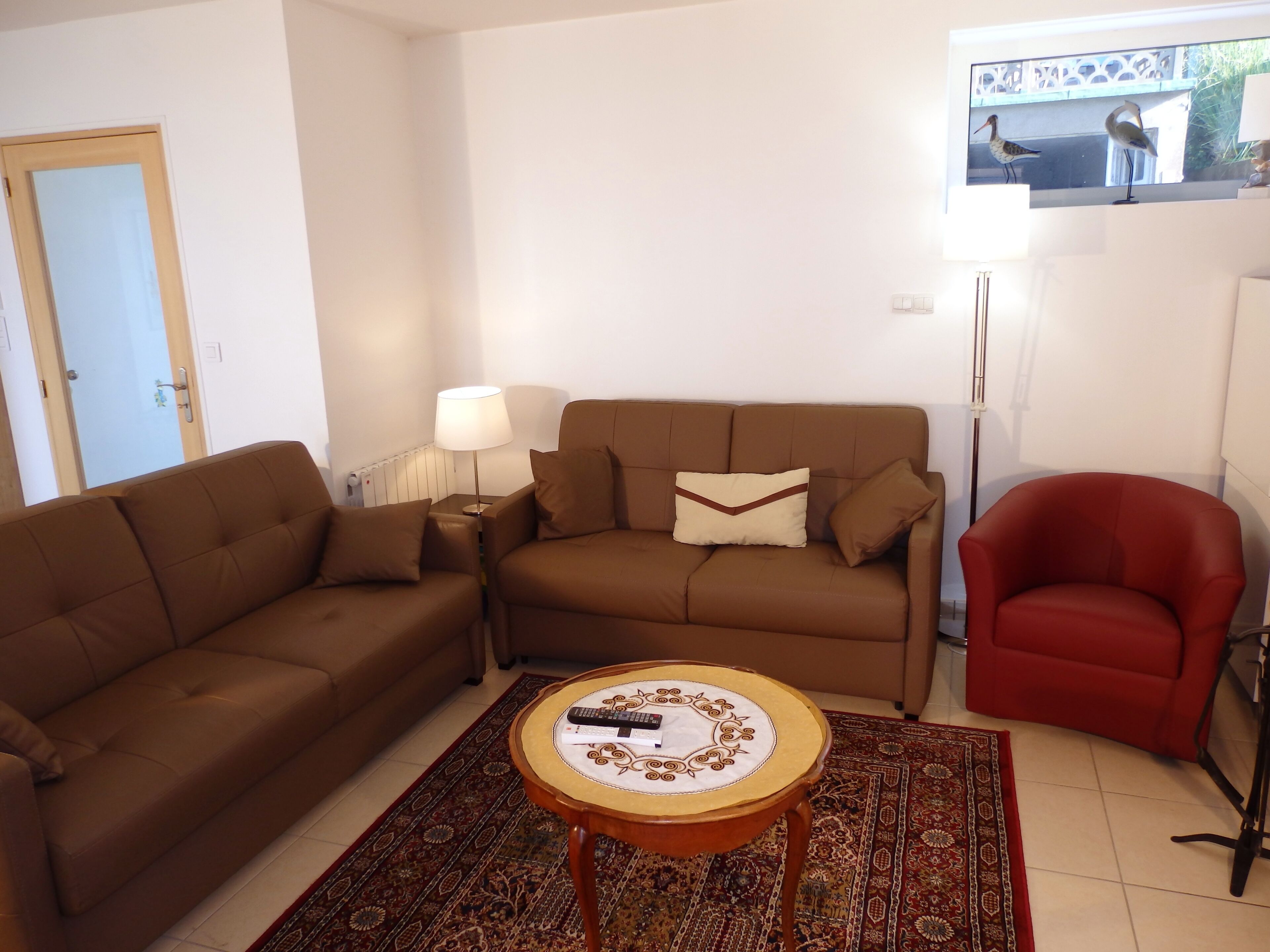 Ein Wohnzimmer mit braunem Sofa, einem roten Sessel, einem Couchtisch mit Fernbedienung und einem gemusterten Teppich.