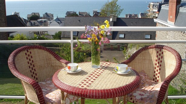 A balcony with wicker chairs and a table, overlooking a coastal view with buildings and the sea. - Modern house with garden and beautiful sea view (Pléneuf-Val-André)