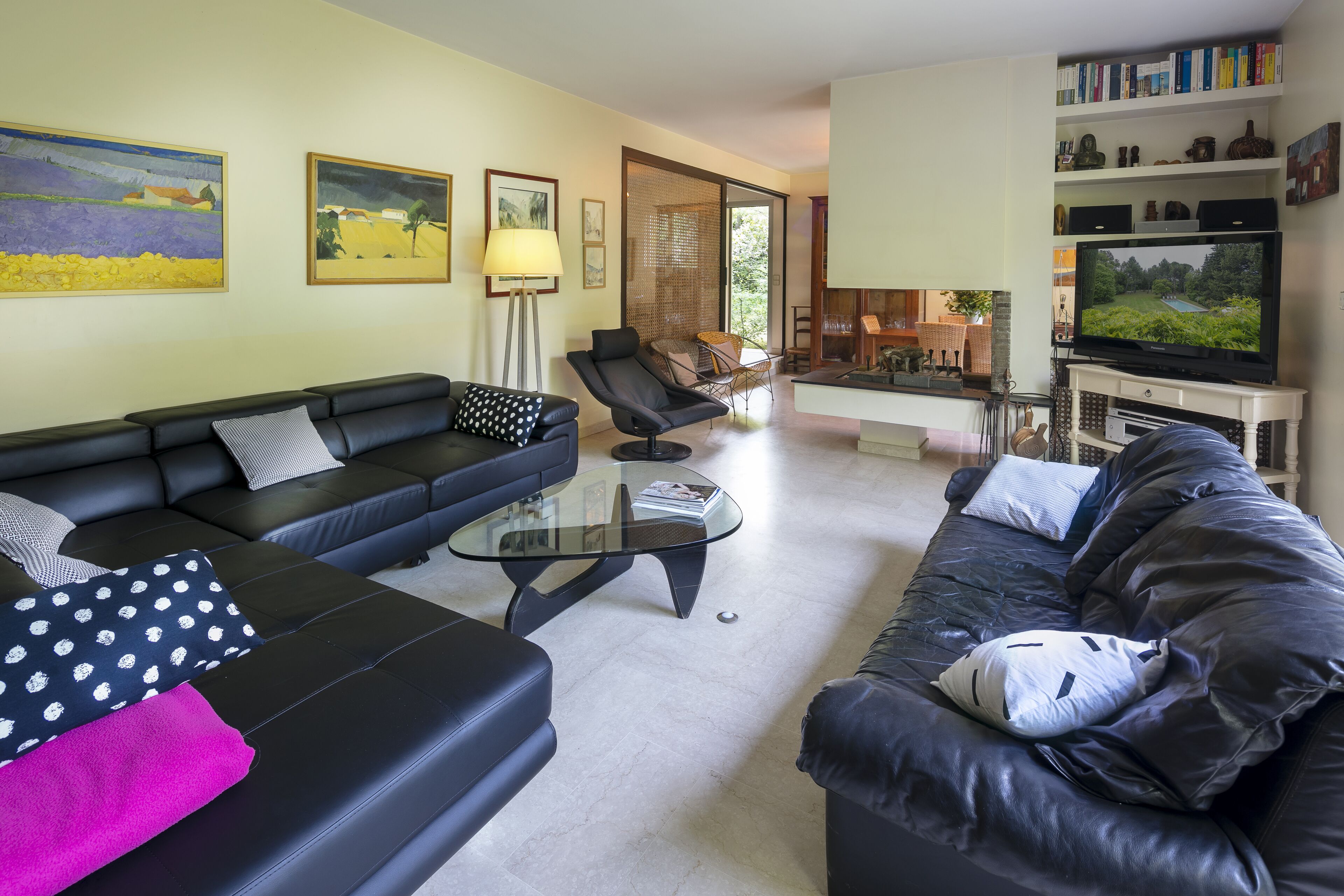 Ein Wohnzimmer mit einer schwarzen Ledersofa, einem Glastisch und einem Fernseher auf einem Schrank. An den Wänden befinden sich Gemälde und ein Bücherregal mit Büchern und Dekoration.
