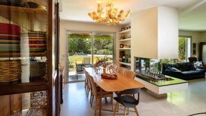 Un comedor moderno con una mesa y sillas de madera, una araña y vistas al exterior a través de amplios ventanales.