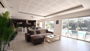 A modern living room with a sofa, ottoman, and coffee table, a kitchen in the background, and a view of a swimming pool through large windows.