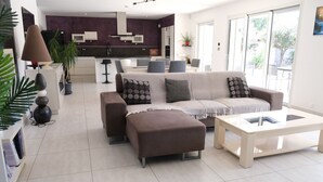 A modern living room with a sofa, ottoman, and coffee table. There is a kitchen in the background with a purple accent wall.